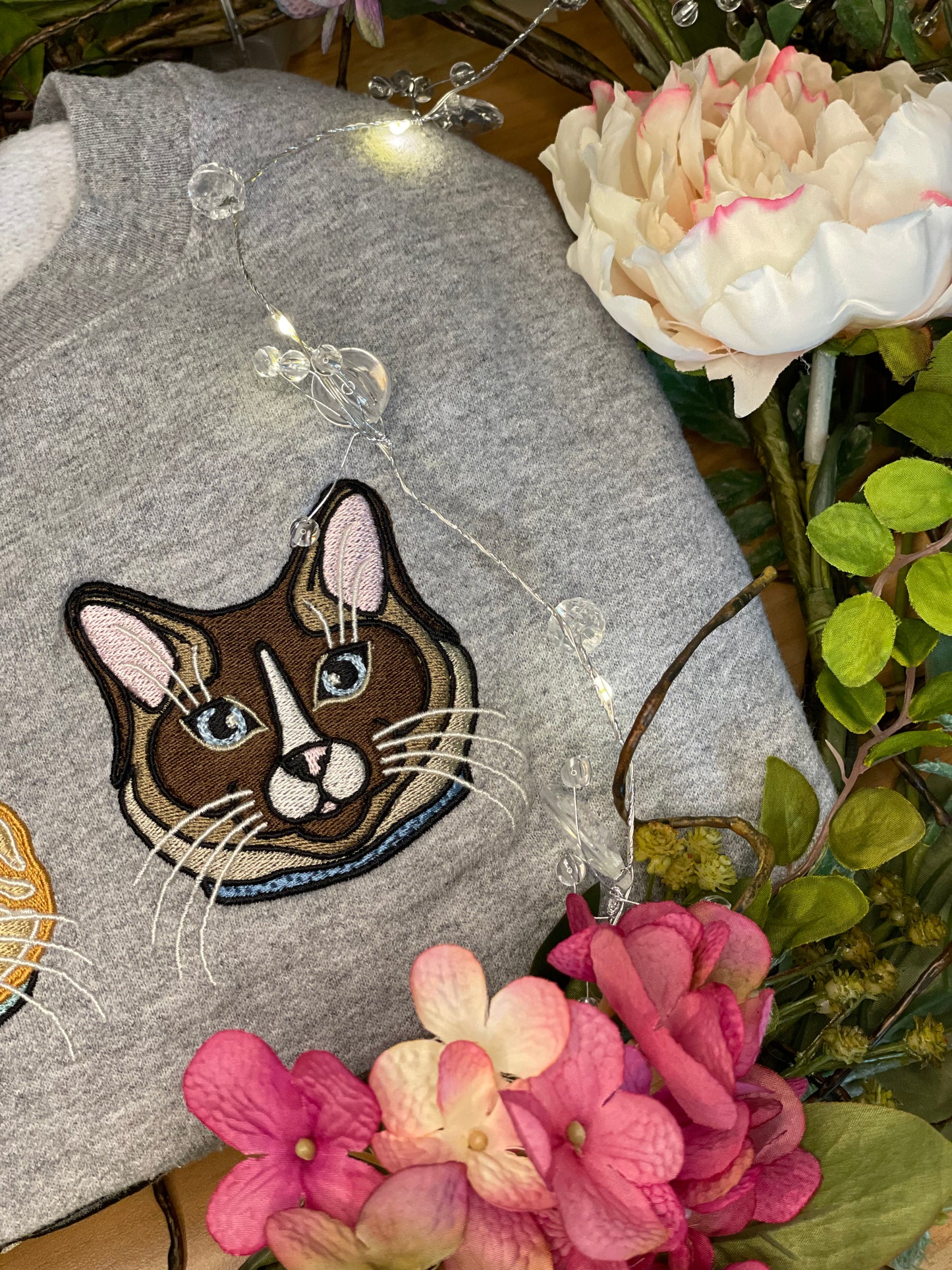 Ragdoll, Snowshoe, Siamese Cat embroidered on a crewneck sweatshirt available in colors baby blue, pigment pink, and forest green design is 4x4 inches on the left chest. Available in sizes S-3XL. Shown on a grey sweatshirt surrounded by flowers. 